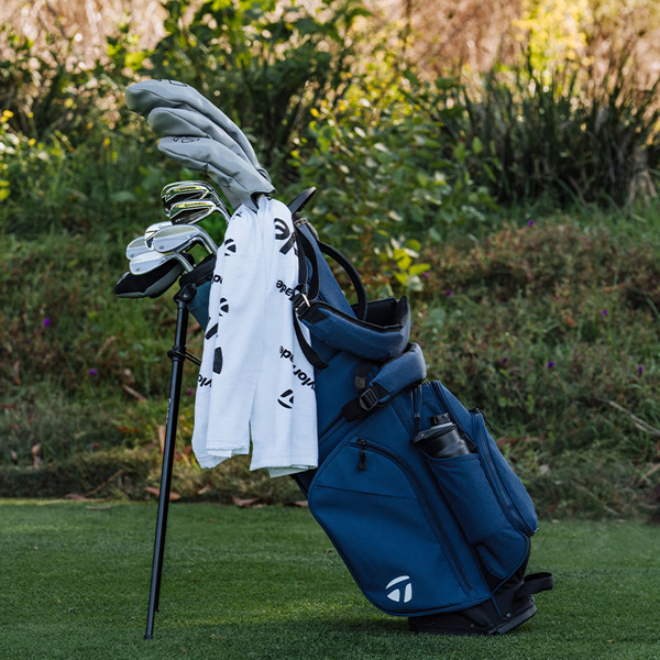 Navy blue TaylorMade Flextech golf bag, outside on the green with plants in the background, clubs in the bag and a TaylorMade towel attached.