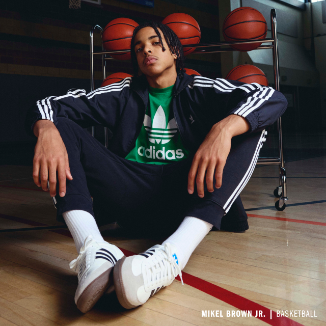 Mikel Brown Jr. sitting on a basketball court wearing an adidas tracksuit, shirt, and sneakers.