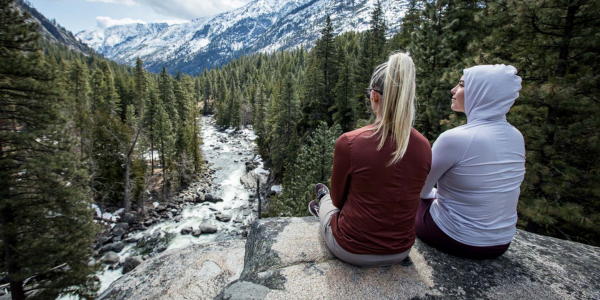 Two women wearing Geo-T's sitting on a rocky ledge overlooking a forested river and snowy mountains in the background