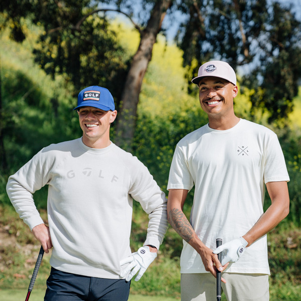 Two golfers smiling on course- one is wearing the blue TaylorMade sunset golf snapback hat and the Golf crewneck in stone heather and the other is wearing the pink TaylorMade Carlsbad 5 Panel snapback hat and the Golf cross t-shirt in sand