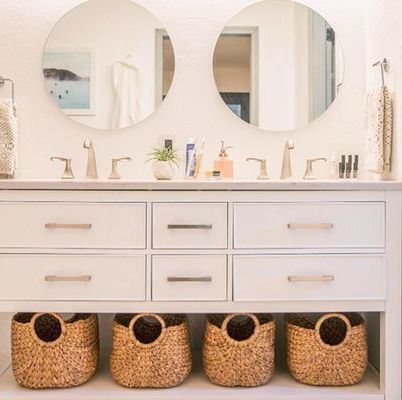 two round mirrors hanging on the wall over a double vanity with gold fixtures and four jute baskets underneath