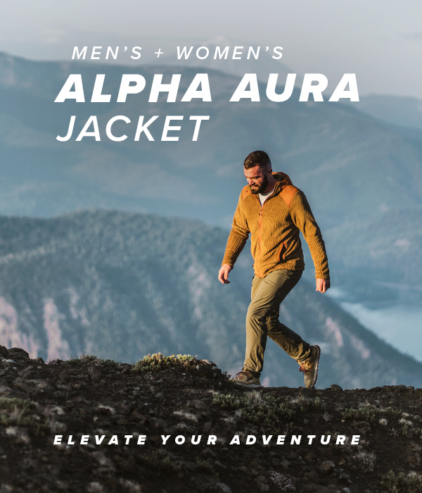 Man wearing the Tobacco colored Alpha Aura jacket while hiking along a ridge line with a mountain lake in the background. Text Overlay saysing "Men's + Women's Alpha Aura Jacket, Elevate Your Adventure"