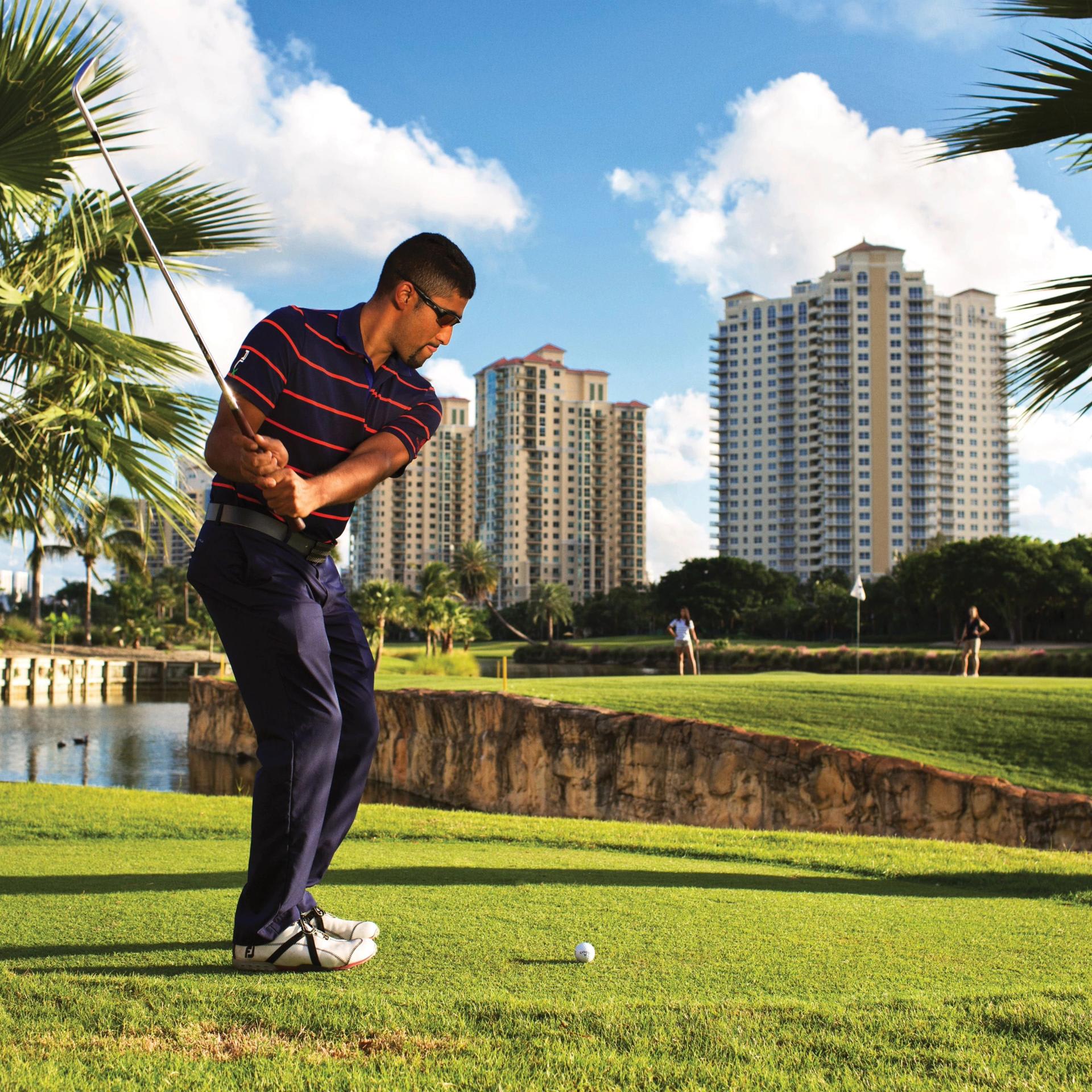 A man gets ready to swing a golf club on a golf course with palm trees, water and buildings in the distance