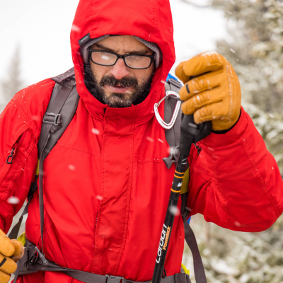 Man wearing a Red Cetra Jacket and Timber Guide Gloves while using trekking poles hiking through the snow.