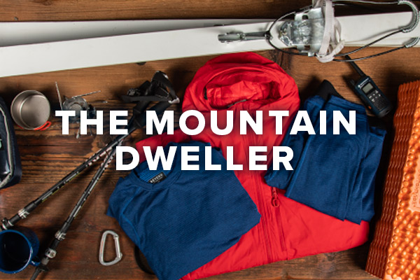 A lay down image of items that a hardened outdoorsman would use on a long excursion in the mountains or other harsh environment. Items include a pair of NATO issued skis, base layers, sleeping pad, insulated jacket, trekking poles and a walkie talkie. Text overlay saying "The Mountain Dweller".