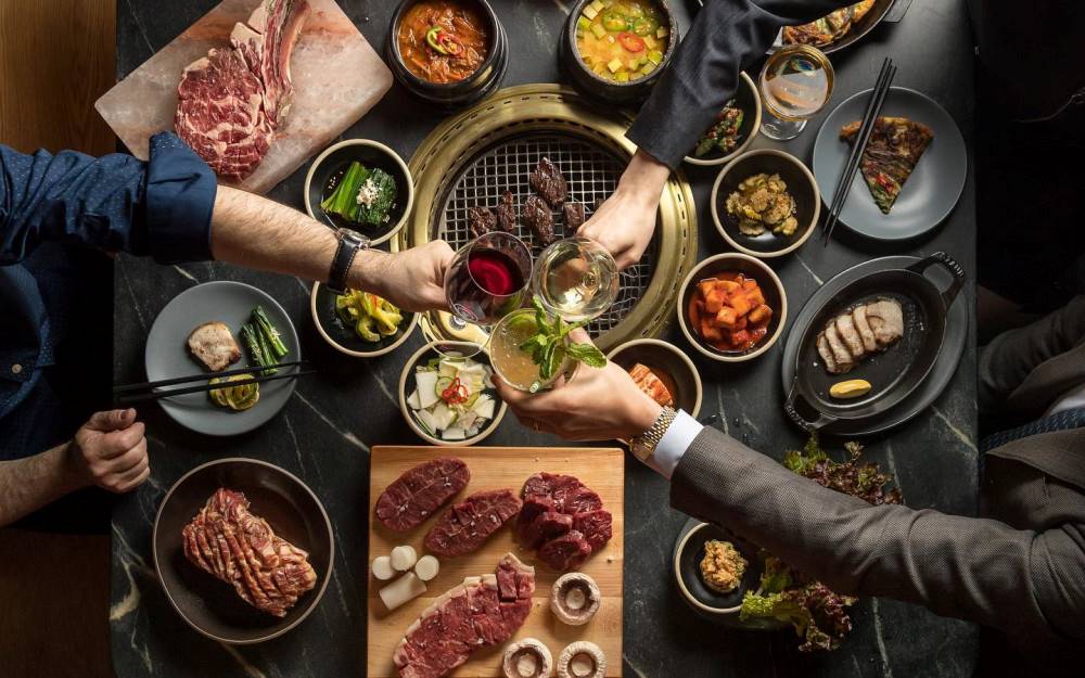 Several hands toast with cocktails over a large dinner spread of meats and vegetables