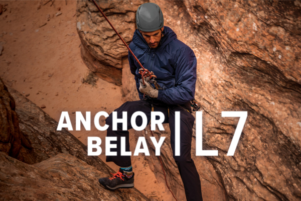 Man wearing the Navy Anchor Belay jacket while rappelling down a cliff side with a text overlay saying "Anchor Belay L7"