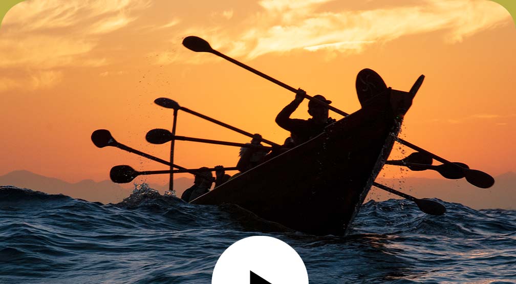 Video play button. Sihouettes of people paddling a canoe on the ocean under orange-colored skies.