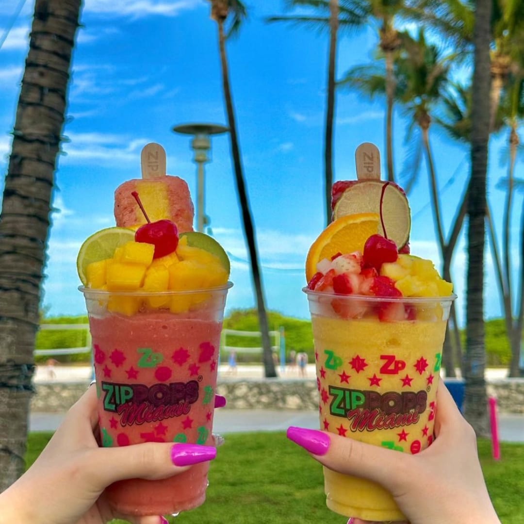 Hands holding two tropical beverages with the beach in the background