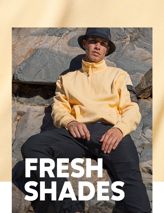 A man taking a break from hiking. He sits on a pile of boulders wearing a yellow fleece and bucket hat. FRESH SHADES overlays the image.