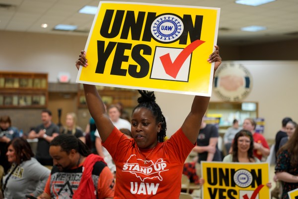 A woman in a red UAW t-shirt holds a yellow sign reading "Union Yes" over her head