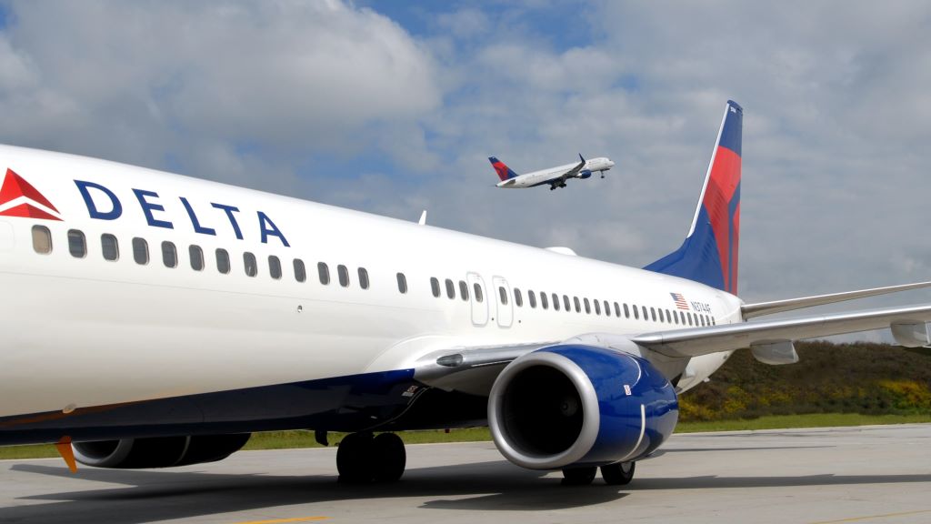 A Delta 737-800 aircraft parked with a Delta plane taking off in the background