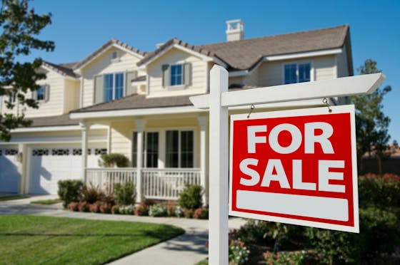 A "For Sale" sign hangs outside of a home with yellow siding