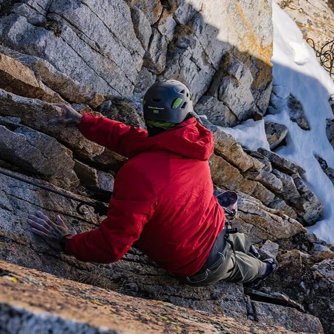 Man rock climbing in the Red Anchor Belay jacket while looking back down the cliff. Snow in the background