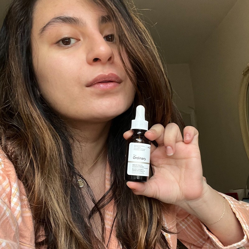 A brown bottle of The Ordinary's Salicylic Acid 2% Anhydrous Solution on a white background (left); A woman with long black hair and a pink long sleeve shirt holding the brown bottle (right)