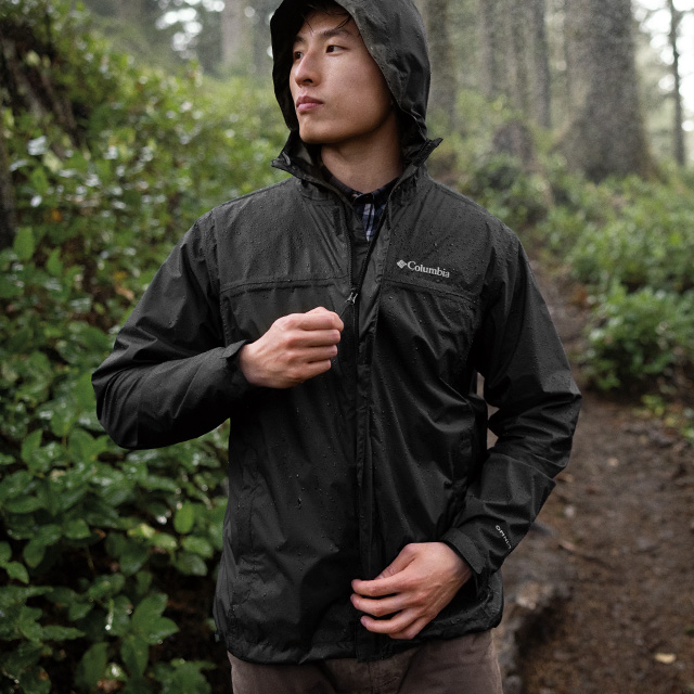 A man on the trail zips up his waterproof jacket with a lush forest behind him.