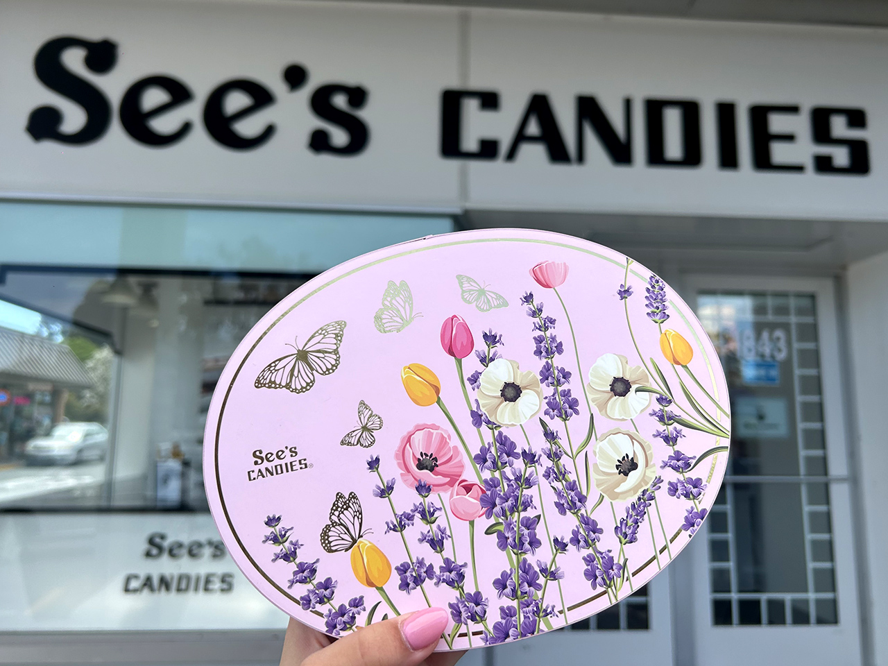 A Hand Holding Up a Spring Bloom Box of Chocolates in Front of a See’s Candies Shop