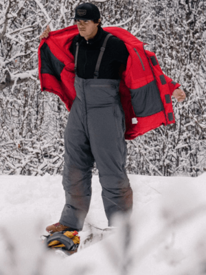 Man wearing the Grey Allta Bibs in the snow with a forest background. He is also putting on the Allta parka in Red.