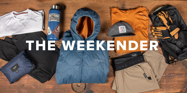 A lay down image of a bunch of Beyond Clothing Products that someone would use on a Weekend outdoor excursion such as a hiking trip. Includes jackets, pants, hats, gloves, shirts. Text Overlay saying "The Weekender".