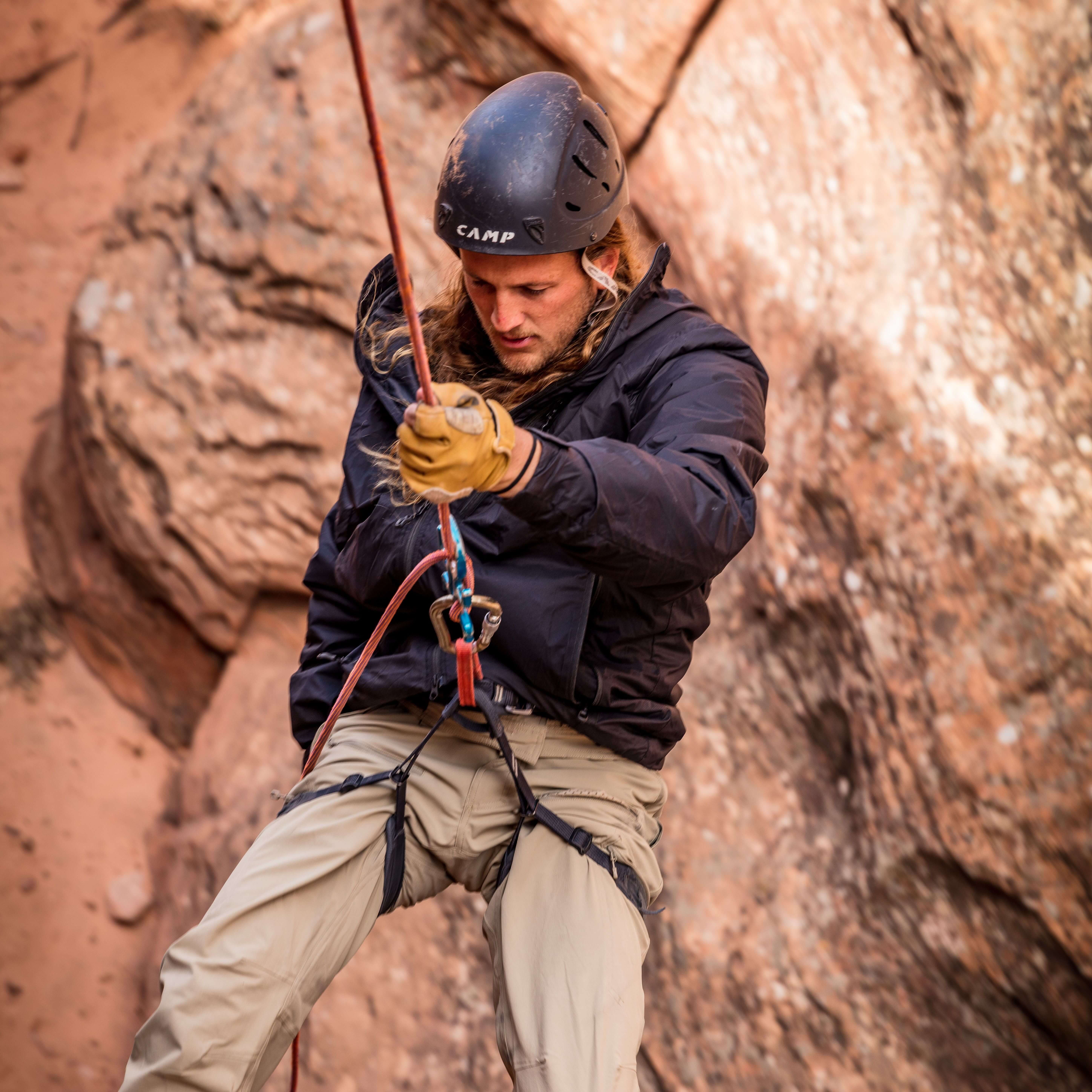 Man rappelling down a cliff side with a rocky background while wearing the Black Anchor Belay jacket