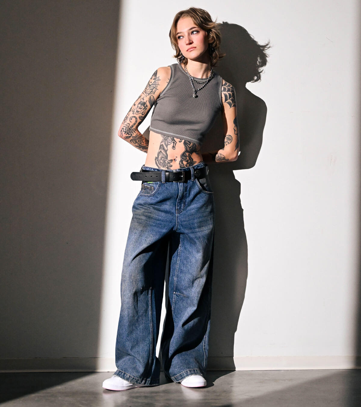 A women leaning on a wall in a cropped gray tank-top, wide jeans, and white shoes.