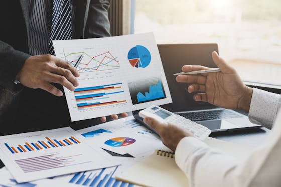 Two men discuss investment strategy over a desk filled with graphs, a computer and a calculator