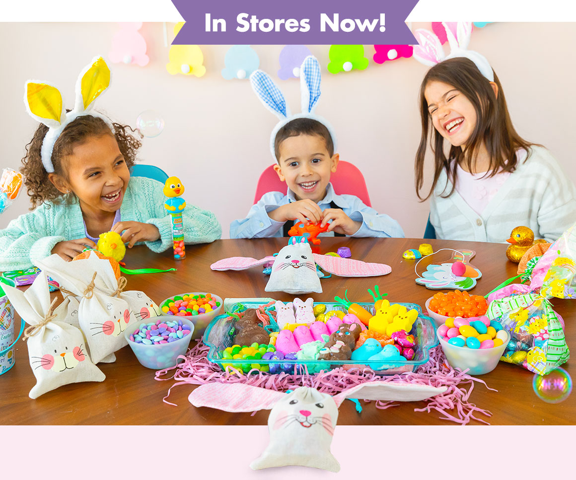 3 children wearing bunny ears sitting at a table with Easter goodie bags, an Easter candy tray, and more